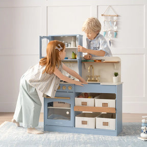 Two children playing with a blue wooden play kitchen set, opening cabinets and interacting with accessories.